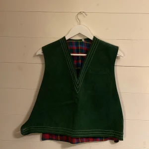 Grön väst i mocka - Cool grön väst i mocka med rödrutigt mönster på insida💚 På utsidan kan man stänga den med knappar på vardera sida och den har vita detaljer vid kragen. Köpt vintage men tyvärr inte kommit till användning.