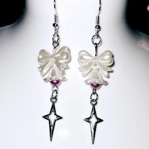 Vit klocka med lila  - Vita akrylpärlor med vit akrylklocka med lila akrylpärla i och avlång silvrig stjärna. Silvriga örhängen (ej nickelfria, ej äkta silver) 