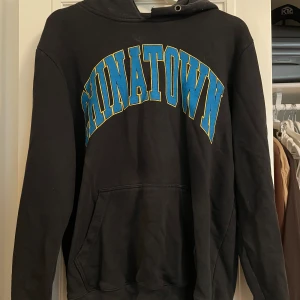Svart hoodie från Chinatown Market - En svart hoodie från Chinatown Market med texten 'CHINATOWN' i blått och gult på framsidan. Hoodien har en stor ficka på magen och en justerbar huva.