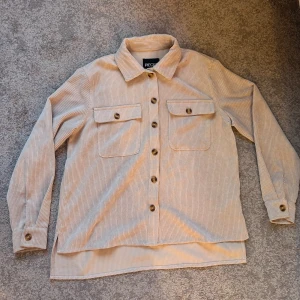 Manchesterskjorta - Beige manchesterskjorta från Pieces. Endast använd 3-4 gånger så den är i väldigt bra skick. Ganska tunn i materialet. Måtten är från kragen till nedersta kanten 56cm, nedersta kanten är 57cm och ärm till ärm 135cm. 