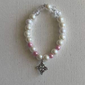 Ett egengjort pärlarmband med färgerna vit och rosa i olika nyanser med en blandning pärlsorter. Armbandet har en berlock i silver som avbildar ett mönster. 