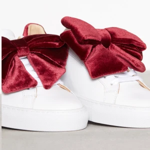 Sneakers med rosett (avtagbar) - Vita sneakers, röda rosetter i sammet, går att ta av.  Köpta på Nelly.com Nypris: 1799kr Nedsatta till 899kr nu men slutsålda. Använda fåtal gånger. Materialet är konst-skinn. Går att putsa till dom renare än på bilden.
