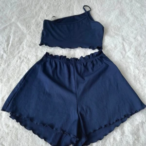 Mörkblå set med topp och shorts - Ett mörkblått set bestående av en ärmlös topp och matchande shorts. Toppen har en enkel design med smala axelband och en rak halsringning. Shortsen har en elastisk midja och vågiga kanter.