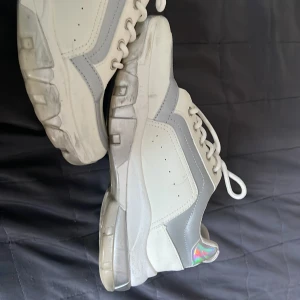 Vita och grå sneakers med holografiska detaljer - Ett par vita och grå sneakers med snörning och holografiska detaljer på hälen. Skorna har en tjock sula och perforerade detaljer på ovansidan för ventilation.