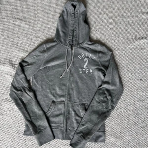Grå hoodie från Hollister - En grå hoodie från Hollister med dragkedja och två fickor framtill. Hoodien har en justerbar. Märket 'Hollister' är tryckt på bröstet. Köpt secondhand. Dem har slitage vid sömmarna o en slags fläck och hål i fickan. Skriv för fler bilder eller frågor! 💚🩶