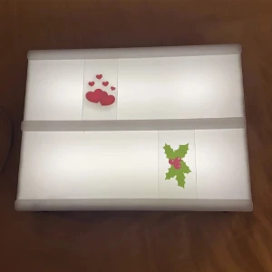 Vit väska med juldekorationer - En vit väska dekorerad med julmotiv, inklusive röda hjärtan och gröna mistelblad med röda bär. Väskan har en rektangulär form och verkar vara tillverkad av ett syntetiskt material.