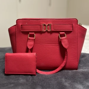 Rosa väska och plånbok - Har använts och kan ha små märken om man kollar väldigt nära och noga, men är i helhet i bra skick. Väskan har två mindre fack i och en dragkedja som förslutning. Plånboken har plats för kort och andra småsaker. Väskan kommer även med ett längre axelband.