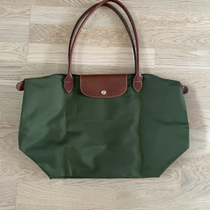 Äkta Longchamp Le Pliage Original M Handbag - Äkta Longchamp väska i modellen Le Pliage Original i storlek M. Äldre modell som inte längre tillverkas. Slitage enligt bilder, ”kantstött”. Lädret är i mycket fint skick. Kontakta för fler bilder. Nypris: 1 499 kr