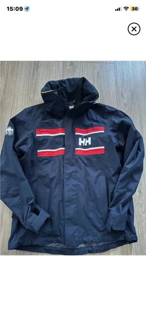 Marinblå Helly Hansen vindjacka M - Snygg marinblå vindjacka från Helly Hansen med röda och vita ränder samt HH-logga fram och Helly Hansen-tryck bak. Jackan har huva, meshfoder och är tillverkad i vattentätt och andningsbart material. Perfekt för blåsiga dagar och outdoor.