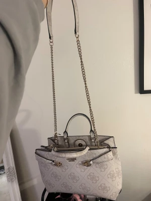 Guess beige handväska med kedja - Snygg beige handväska från Guess med logomönster och guldfärgade detaljer. Väskan har både handtag och en lång axelrem med kedja, samt flera fack och dragkedja inuti. Perfekt storlek för allt du behöver och riktigt stilren design. Helt ny och oanvänd säljs för 1700kr men pris kan diskuteras vid intresse.