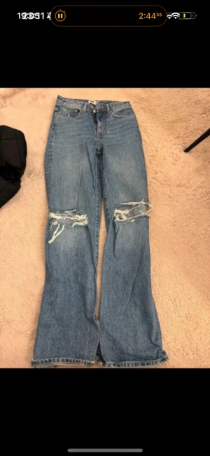 Blå ripped jeans med vida ben - Säljer ett par blå jeans med höga knäslitsar och slitna detaljer över knäna. Byxorna har en klassisk femficksmodell, hög midja och vida ben. Perfekta för en avslappnad och trendig look.