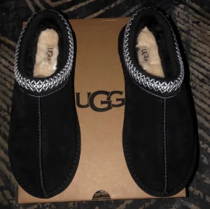 Svarta UGG tofflor i mocka - Mysiga svarta UGG tofflor med mjukt fårskinnsfoder och ovandel i mocka. Snyggt mönstrad kant i vitt och svart runt öppningen. Perfekta för att hålla fötterna varma hemma. Platt sula med UGG-logga undertill.