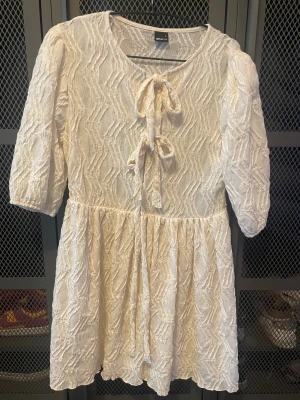 Vit kortklänning med knyt från Gina Tricot - Supersöt vit kortklänning från Gina Tricot med strukturmönster och två knyt framtill. Klänningen har halv-långa puffärmar och en flowy kjol. Perfekt för sommarens alla tillfällen och ger en romantisk vibe.