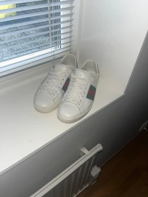Gucci vita sneakers med röd/grön rand - Säljer ett par fräscha vita sneakers från Gucci med klassiska röd och grön rand på sidan. Skorna har snörning, rund tå och platt sula. Tillverkade i skinn för en lyxig känsla och passar perfekt till jeans eller streetwear. Kan gå ner i pris vid snabbt köp!