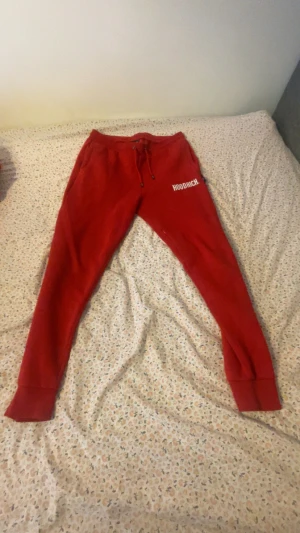 Röda mjukisbyxor från Hoodrich - Snygga röda mjukisbyxor från Hoodrich med vit logotyp på vänster lår. Byxorna har snörning i midjan och ribbade muddar vid bensluten. Perfekta för chill eller träning, med en slim passform och mjukt bomullsmaterial.