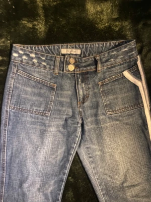 Blå jeans med vita detaljer från beyond retro original pris 400 - Snygga blå jeans från beyond retro med raka ben och coola vita detaljer längs sidorna och på bakfickan. Jeansen har framfickor med markerade sömmar och dubbla knappar i midjan. Perfekt för dig som gillar unik stil och streetvibe.