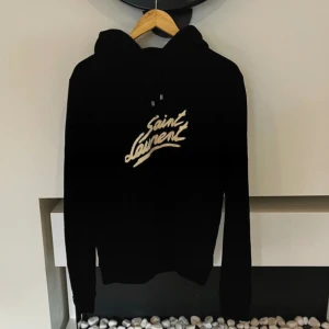  Saint Laurent hoodie  - Svart hoodie från Saint Laurent med stort vitt logotryck på bröstet. Tröjan har huva med snörning och långa ärmar. Perfekt för dig som gillar streetstyle och vill ha en ikonisk look. Materialet känns mjukt och bekvämt mot huden.