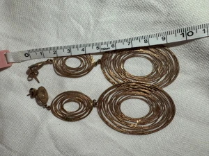 Set med stora guldiga/rose örhängen och armband - Säljer ett matchande set med stora statement-örhängen och ett armband i guldtonad metall. Båda har coola cirkel- och ovalformer med flera ringar i lager, vilket ger en unik och trendig look. Perfekt för dig som gillar att sticka ut med accessoarerna.