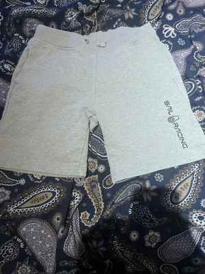 Grå Sail Racing shorts med tryck - Snygga grå shorts från Sail Racing med svart logotyptryck på ena benet. Modellen har ribbad midja med knapp och hällor, samt fickor på sidorna. Mjukt material som känns skönt mot huden, perfekt för chill dagar eller sport. Passar dig som gillar enkel och clean stil.