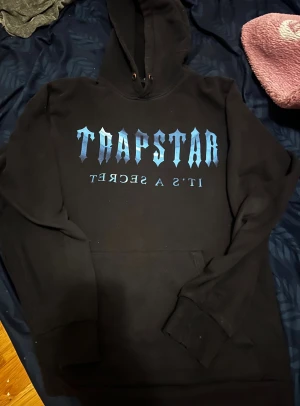 Svart hoodie Trapstar med blått tryck - Svart hoodie från Trapstar med stort blått tryck på bröstet där det står 'TRAPSTAR IT'S A SECRET'. Tröjan har huva med snörning och en klassisk magficka. Tillverkad i 100% bomull, perfekt för dig som gillar streetwear och vill sticka ut.