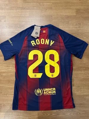 FC Barcelona hemmatröja #Roony28 - Fc Barcelona hemmaställ för säsongen 25/26 med Roony 28 på ryggen. Tröjan är helt ny och tillgänglig i storlek M. Bästa kvalitet som alltid!!