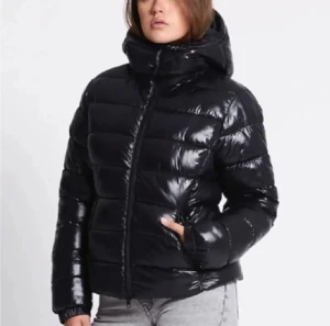 Svart glansig pufferjacka med huva - Svart pufferjacka med glansig finish och stor huva. Jackan har dragkedja framtill, knappar vid kragen och breda, quiltade paneler. Perfekt för kalla dagar och riktigt snygg till jeans eller cargobyxor.