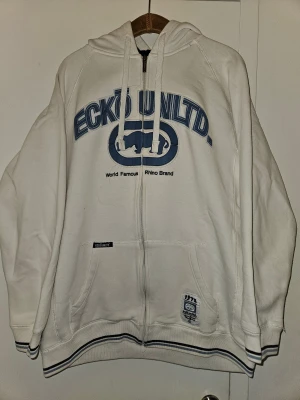 Ecko Unltd hoodie - Vit hoodie från Ecko Unltd. i storlek L med blått tryck och rhino-logga på bröstet. Har liten fläck på ryggen