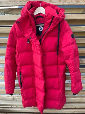 Röd dunjacka från Reset  - Snygg röd dunjacka från Reset med huva och svarta detaljer. Jackan är quiltad, har dragkedja och knappar framtill samt praktiska fickor med dragkedja. Perfekt för kalla dagar när du vill hålla stilen och värmen.