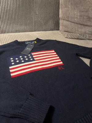 Mörkblå stickad tröja Polo Ralph Lauren - USA - Mörkblå stickad tröja från Polo Ralph Lauren med stor amerikansk flagga på bröstet och RL-broderi i rött. Tröjan har rund hals, ribbade muddar och är långärmad. Perfekt för dig som gillar klassisk och cool stil. Väldigt liten i storleken Längd 61,5 cm