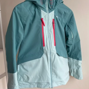 Skidset med jacka och byxor  - Snygg tvåfärgad skidjacka i blå och ljusblå nyanser med coola rosa detaljer. Matchande rosa skidbyxor med RECCO-reflektor. Jackan har huva, hel dragkedja och snölås. Skidsetet är vattentätt (10 000 vattenpelare). Perfekt för skidbacken och andra utomhusaktiviteter. Toppenkvalitet och mycket bra skick, endast använts ett fåtal gånger.