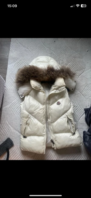 Vit dunväst med päls Moncler - Exklusiv vit dunväst från Moncler med stor huva och lyxig pälsdetalj. Västen har dragkedja framtill, två fickor med dragkedja och broderad Moncler-logga på bröstet. Bred resår med Moncler-text baktill ger en snygg siluett.