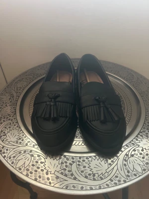 Svarta loafers med tofsar - Stilrena aldrig andvända svarta loafers i skinnimitation med dekorativa tofsar och fransdetaljer framtill. Skorna har en klassisk rund tå och platt sula, perfekt för en snygg och chill look. Passar till både jeans och kostymbyxor.