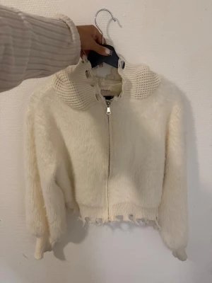 Vit stickad kofta från LTT Studio - Säljer en vit stickad kofta från LTT Studio med dragkedja framtill och grovstickad krage. Koftan har långärmade ärmar och en cool, ruffad kant nedtill och vid kragen som ger en edgy vibe. Perfekt för dig som gillar unika detaljer och mjuka material.