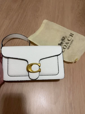 Vit handväska från Coach med gulddetaljer - Snygg vit handväska från Coach i strukturerat skinn med guldfärgat C-spänne framtill. Väskan har svarta kanter, avtagbar axelrem och bredare handtag. Perfekt storlek för det viktigaste och stilren design som lyfter din look.