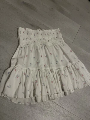 Vit blommig kjol - Supersöt kort kjol från lager 157 i vitt med små blommor i rosa och blått. Kjolen har smock i midjan och volanger nertill för en flowy känsla. Perfekt för dig som gillar romantisk och feminin stil. Passar XS/S