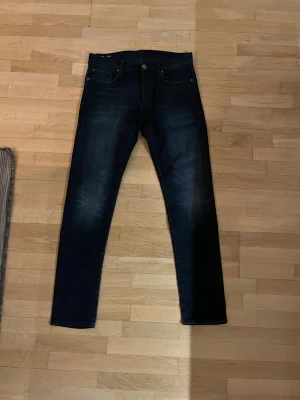 G-star jeans slim fit 30/34 - Snygga svart/mörkblå jeans från G-Star RAW i storlek 30/34. Modellen har klassisk femficksdesign, slimmade ben och en snygg wash. Jeansen är tillverkade i ett denimtyg som sitter skönt och följer kroppen. Perfekt för dig som gillar stilrena och moderna jeans.