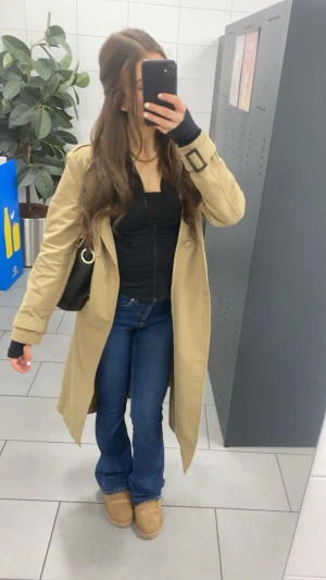 Beige trenchcoat med bälte - Stilren beige trenchcoat med klassisk krage och bälte i midjan. Kappan har långa ärmar med spänne vid ärmslut och knäppning framtill. Perfekt för lager-på-lager och ger en tidlös look till din outfit.