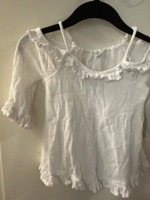 Vit volangblus från Lindex strl: 128 - Supersöt vit blus från Lindex  i storlek 128. Blusen har trekvartsärmar, volangdetaljer vid halsringning, ärmslut och nederkant samt tunna axelband. Tillverkad i ett lätt, krinklat bomullstyg som ger en luftig känsla.