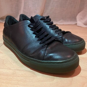 Svarta sneakers från Axel Arigato - Svarta sneakers från Axel Arigato med stilren design och gröna räffliga sulor. Skorna är tillverkade i skinn och har svarta snören samt rund tå. Perfekta för dig som gillar minimalistisk och clean stil.