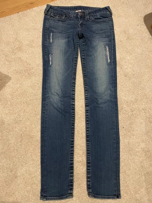 True Religion blå bootcut jeans, stl 29 - Snygga blå jeans från True Religion med slitna detaljer framtill. Klassiska bakfickor med dekorativa sömmar och knappar. Började sy om jeansen så det är som en slits går enkelt att sy tillbaka eller sy om. Perfekt för dig som gillar en avslappnad men trendig look.