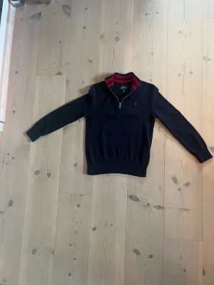 Mörkblå half zip tröja Polo Ralph Lauren - Snygg mörkblå stickad tröja från Polo Ralph Lauren med röd broderad logga på bröstet. Kragen har coola röda ränder och en half zip-dragkedja. Långärmad och ribbad vid ärmslut och nederkant. Perfekt för dig som gillar klassisk stil med en twist.