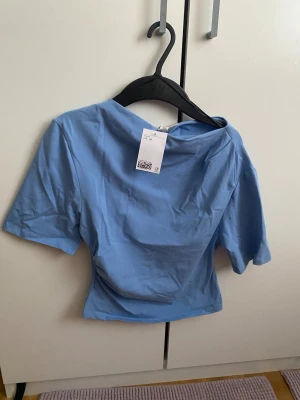 Ljusblå topp från H&M, storlek XS - Säljer en ljusblå topp från H&M i storlek XS. Toppen har korta ärmar och en något croppad passform med mjukt, stretchigt material i bomull och elastan. Enkel och clean design med rundad hals och slät yta, perfekt för att styla med jeans eller kjol.