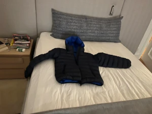 Svart och blå pufferjacka med huva - Säljer en svart pufferjacka med blå insida och huva. Jackan har horisontella quiltade sektioner, dragkedja framtill och två sidofickor med dragkedja. Perfekt för kalla dagar och har en cool kontrastfärg på insidan.