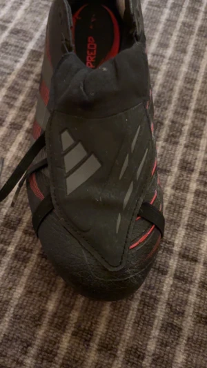 Adidas Predator fotbollsskor svart/röd - Säljer ett par Adidas Predator fotbollsskor i svart med röda och grå detaljer. Skorna har snörning, platt sula med dobbar och en sportig design med mönstrade paneler. Tillverkade i syntetmaterial för bra grepp och kontroll på planen.