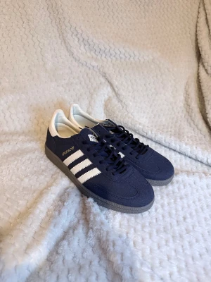 Adidas Spezial sneakers marinblå - Adidas Spezial sneakers i marinblå mocka med klassiska vita ränder och vit häl. Snygg retrostil med grå platt sula och rund tå. Skorna har snörning och guldfärgad Spezial-logga på sidan. Perfekta för dig som gillar streetwear och sportig vibe.