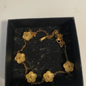 halsband rostfritt stål  - Snyggt guldfärgat halsband med fem blommor i relief på en tunn kedja. Halsbandet är tillverkat i rostfritt stål och har en elegant och trendig design som sticker ut. Perfekt accessoar för att lyfta din outfit. Ask ingår ej
