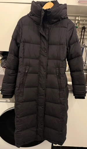 Svart lång dunjacka Rockandblue - Lång svart dunjacka från Rockandblue med hög krage, huva och dragkedjor i sidorna. Jackan har quiltad design, två fickor med dragkedja och ribbade muddar vid ärmsluten. Perfekt för kalla vinterdagar.