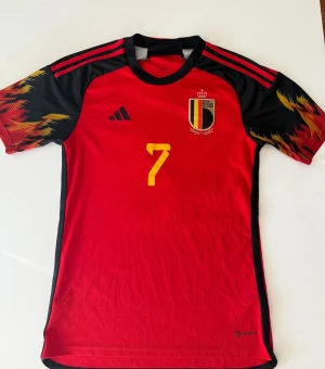 Belgien De Bruyne #7 fotbollströja Adidas - Säljer en officiell Belgien landslagströja från Adidas med De Bruyne och nummer 7 på ryggen. Tröjan är röd med svarta och gula detaljer, korta ärmar med eldliknande mönster, och har landslagets emblem på bröstet. Tillverkad i lätt polyester med Aeroready-teknologi.