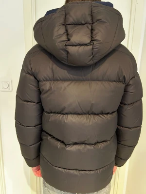 Svart Colmar pufferjacka med huva - Säljer en svart pufferjacka från Colmar med stor huva och två stora fickor framtill. Jackan har en clean look, diskret logga på ärmen och dragkedja hela vägen upp. Fylld med dun för extra värme, perfekt för kalla dagar.