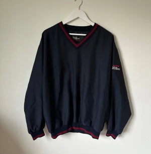 Marinblå v-ringad tröja Oscar Jacobson - Marinblå v-ringad windbreaker tröja från Oscar Jacobson med röda och svarta detaljer vid halsringning och muddar. Långärmad och loose passform, med broderad logga på ärmen. Materialet är syntet och tröjan har ribbade muddar för extra stil. 4Ä11